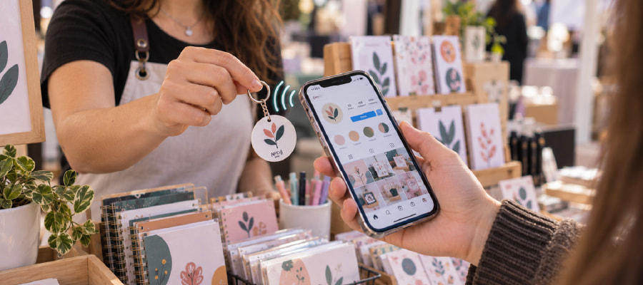 Emprendedora en una feria de papelería muestra un llavero NFC personalizado mientras una persona acerca su celular para acceder al perfil de Instagram de la marca.
